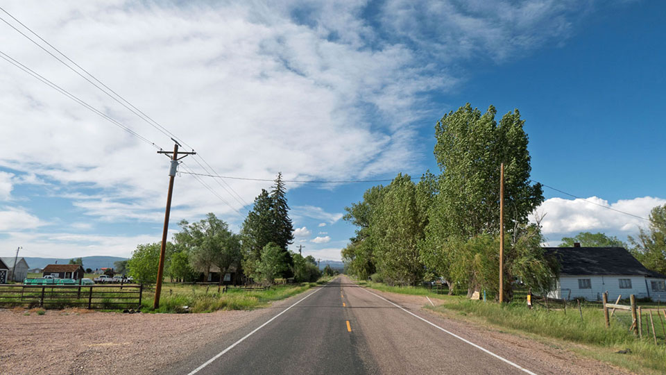 Rural road flanked with large trees, old ranger station on one side and a house on the other under blue sky.