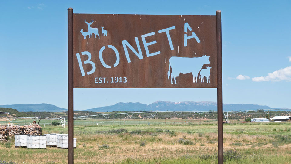 A cut sheet metal sign that says Boneta est. 1915. Mountains and cedar covered hills in the background.