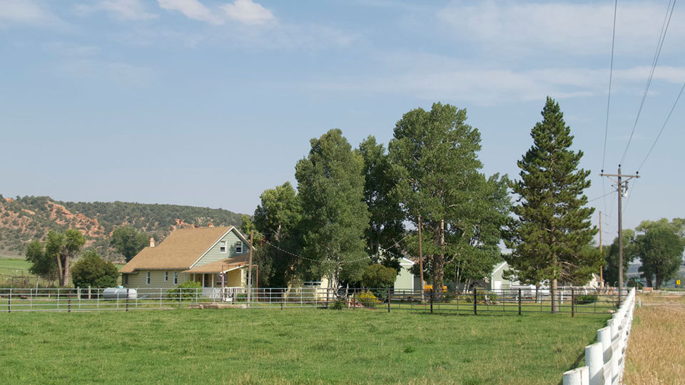 A pale green farmhouse surrounded by green fields and red hills dotted with juniper trees