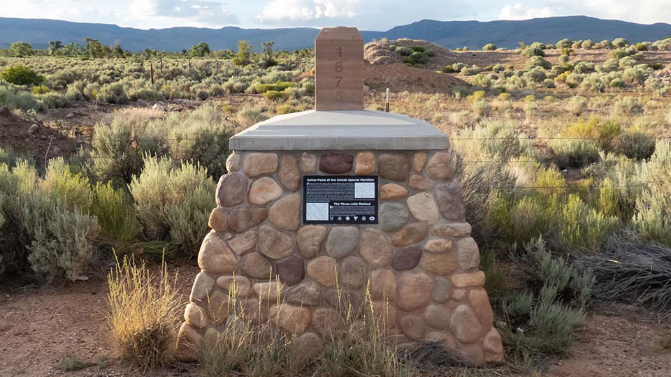 Stone monument marking the initial point of the Uintah Special Meridian, with a wooden marker post reading '1876' and a plaque, set among sagebrush with mountains in the background.
