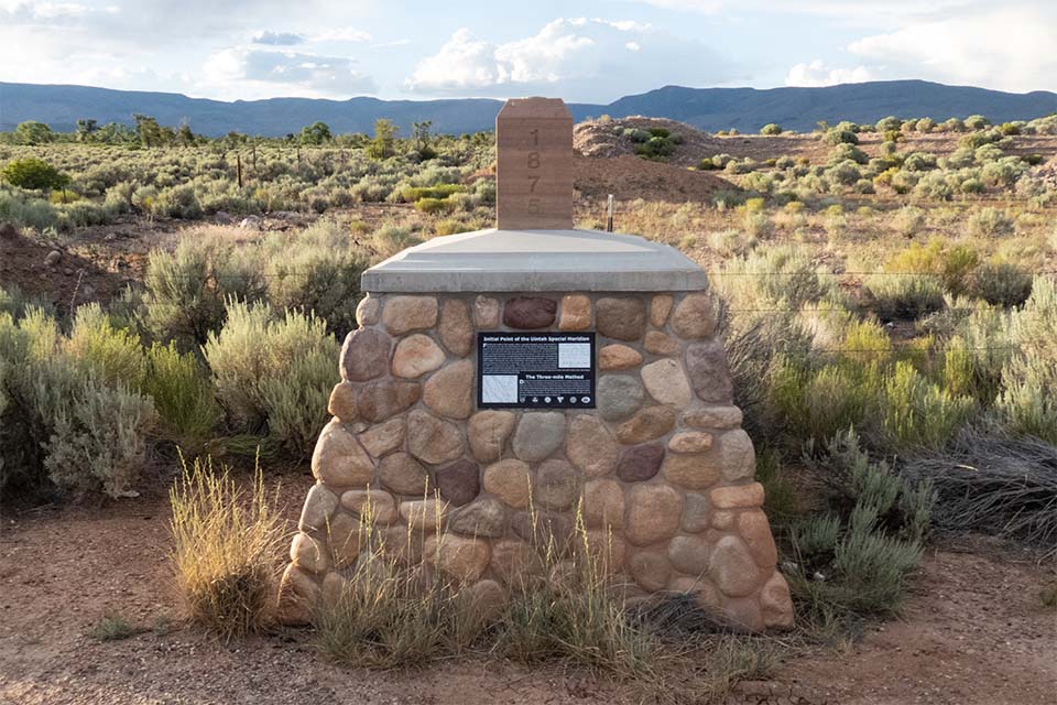 Stone monument marking the initial point of the Uintah Special Meridian, with a wooden marker post reading '1876' and a plaque, set among sagebrush with mountains in the background.
